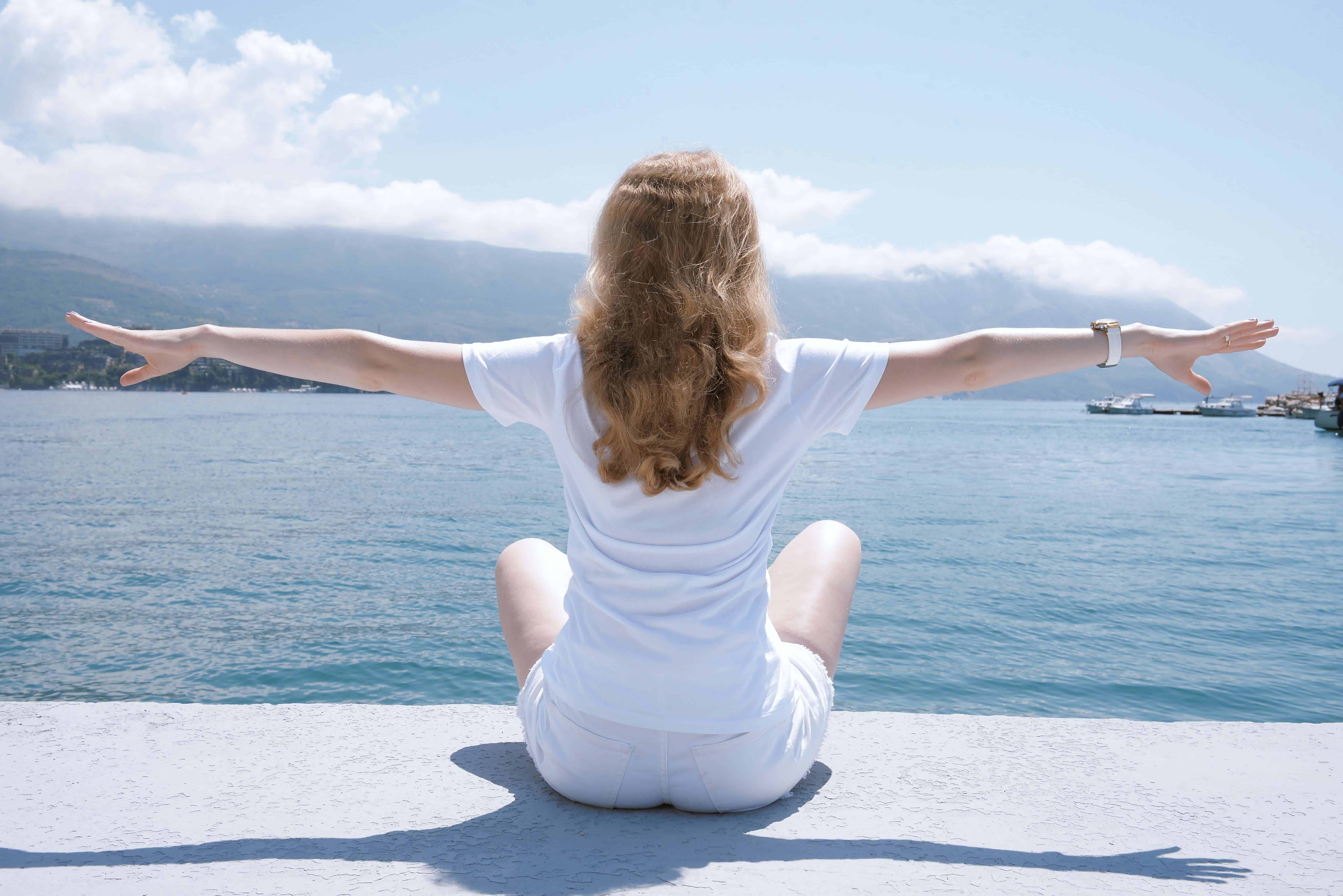 A woman in a white shirt and shorts sitting and facing the ocean with her arms reaching out to the sides. woman in white shirt and shorts residential laundry services